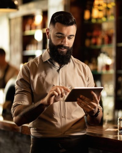 Smiling barista using digital tablet while working in a bar. Young smiling waiter using touchpad while working in a pub.